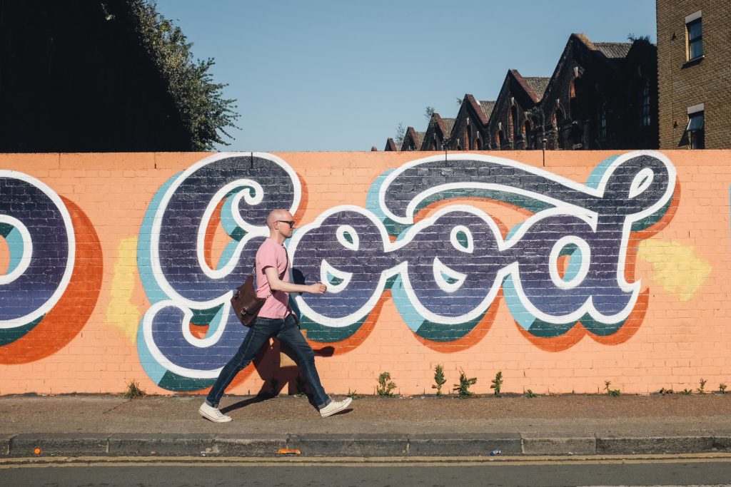 A photo of a man walking past some colourful street art which says the word good in big cursive letters.