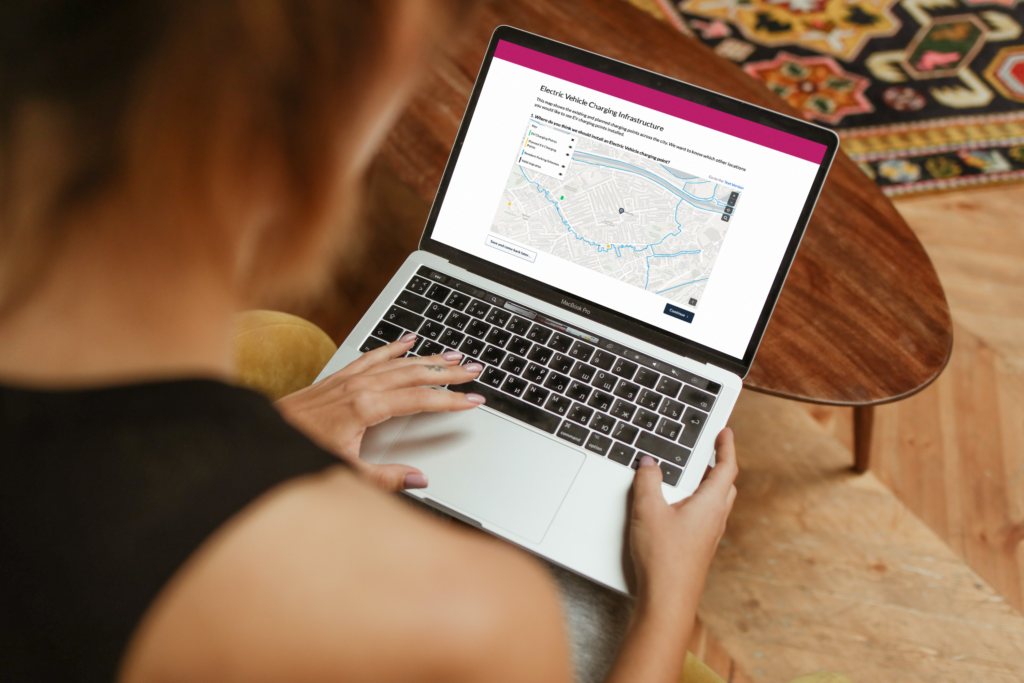 over-shoulder image of woman looking down at a laptop on her lap. the screen displays an Electric Vehicle Charging Infrastructure consultation, using Citizen Space Geospatial's drop a pin activity type
